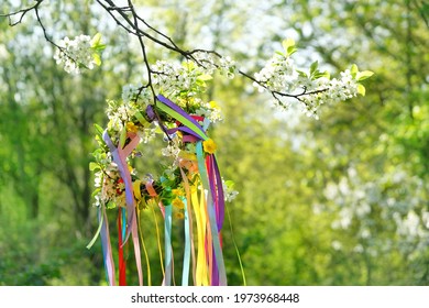Spring flower wreath with colorful ribbons on tree in garden, green natural background. floral decor. Symbol of Beltane, Wiccan Celtic Holiday beginning of summer. pagan witch traditions, rituals