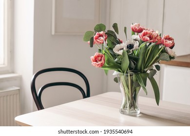 Spring flowers in glass vase on wooden table. Blurred kitchen background with old chair. Bouquet of red tulips, white anemone flowers and eucalyptus branches. Contemporary elegant scandi interior.