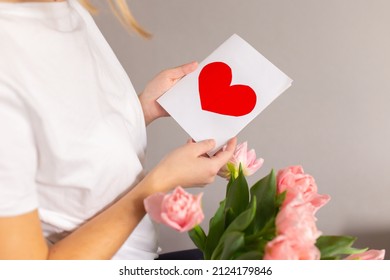 Spring Flowers And Greeting Card. Happy Mom Holds A Card With A Red Heart In Her Hands