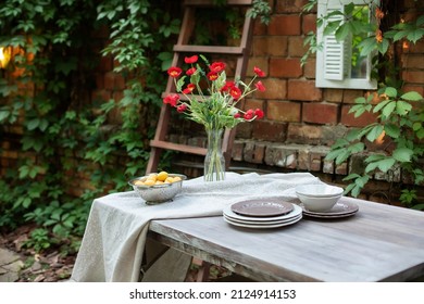 Spring Flowers in a vase at home. Wooden table and plates for garden party or dinner. Table for lunch outside in garden in patio yard of house. Summer bouquet of red poppies in a glass vase on Terrace