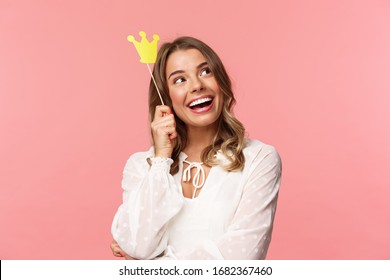 Spring, happiness and celebration concept. Close-up portrait of dreamy beautiful young blond girl imaging something cute and romantic, smiling look up daydreaming with crown on stick, pink background