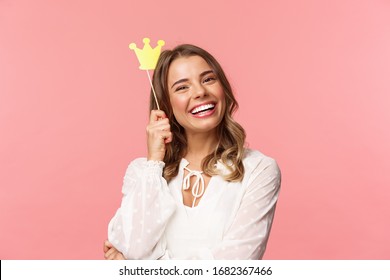 Spring, happiness and celebration concept. Close-up portrait of charming smiling, lovely blond girl holding small queen crown on stick, laughing joyfully, feel empowered and happy, pink background