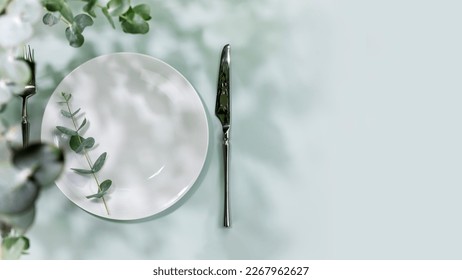 Spring holiday table settings. Empty plate with fork and knife on blue background with eucalyptus leaves. Mockup of romantic summer holiday card with setting suitable for menu. Flat lay, copy space