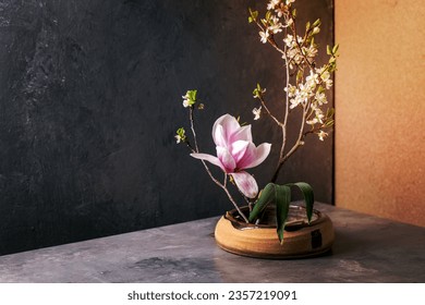 Spring ikebana. Floral composition with spring blooming magnolia and plum branch flowers in brown ceramic bowl, standing on grey table. Japanese style home decor. Copy space