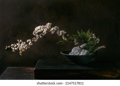 Spring ikebana. Minimalistic floral composition with spring blooming white flowers, thuja branch and stones in black ceramic bowl, standing on black wooden table. Japanese style home decor - Powered by Shutterstock