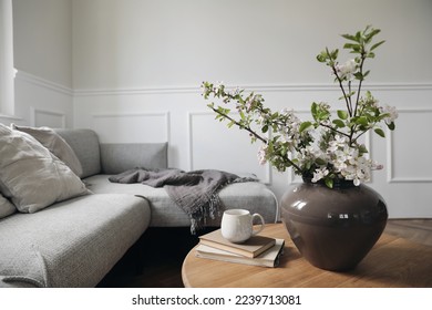 Spring interior still life. Cup of coffee on pile of books. Round wooden table. Blossoming apple tree branches in ceramic vase. Cozy linen sofa with cushions. Elegant living room view. Home decor. - Shutterstock ID 2239713081