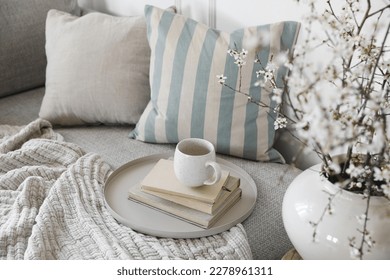 Spring interior still life. Cup of coffee, tea on pile of books. Round beige table. Blossoming cherry plum tree branches in ceramic vase. Cozy linen sofa, cushions. Blurred background. Home decor. Top - Shutterstock ID 2278961311