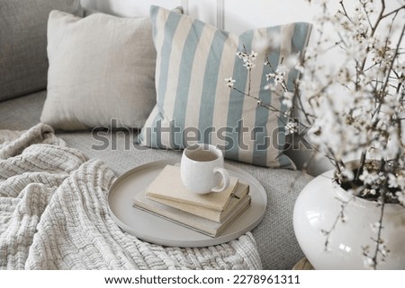Spring interior still life. Cup of coffee, tea on pile of books. Round beige table. Blossoming cherry plum tree branches in ceramic vase. Cozy linen sofa, cushions. Blurred background. Home decor. Top