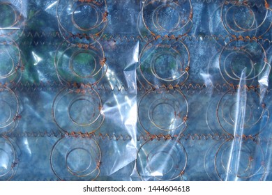 Spring Mattress Wrapped In Plastic Film Wrap Creates A Geometric Texture And A Repetitive Pattern With Its Circular Spring Wires. It Is Thrown Out To The Street As Garbage.