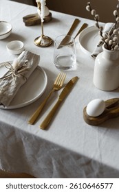 Spring minimalist table setting in warm neutral colors. Easter festive table decoration, white plates, gold cutlery, candles, eggs and willow branches in vase