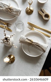 Spring minimalist table setting in warm neutral colors. Easter festive table decoration, white plates, gold cutlery, candles, eggs and willow branches in vase