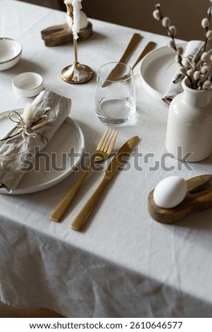 Spring minimalist table setting in warm neutral colors. Easter festive table decoration, white plates, gold cutlery, candles, eggs and willow branches in vase