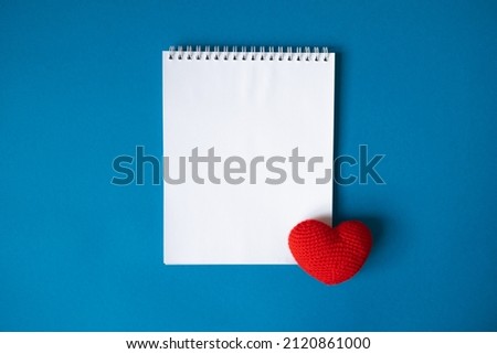 spring notepad. open on a blank white page. a red knitted heart adorns the corner of the page. Blu background