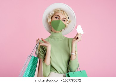 Spring Online Shopping During Quarantine Conception: Fashionable Woman Wearing Protective Mask Posing With Colorful Paper Bags And Plastic Bank Card. Pink Background. Copy, Empty Space For Text