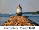 Spring Point Ledge Lighthouse at the end of a granite breakwater with sailboat on Casco Bay in warm golden hour light in South Portland, Maine, USA