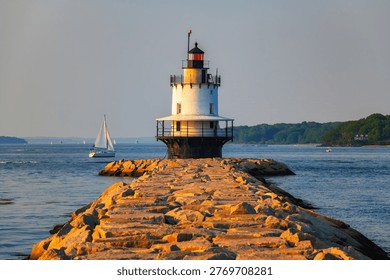 Spring Point Ledge Lighthouse at the end of a granite breakwater with sailboat on Casco Bay in warm golden hour light in South Portland, Maine, USA