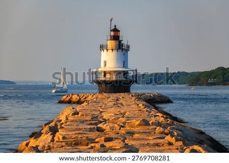 Spring Point Ledge Lighthouse at the end of a granite breakwater with sailboat on Casco Bay in warm golden hour light in South Portland, Maine, USA