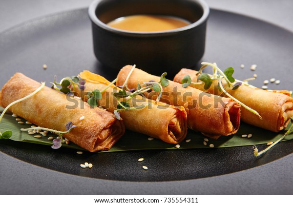 Spring rolls - fried Vietnamese rolls of rice\
edible paper on black matte plate next to cup with sauce. Pan-Asian\
menu of restaurant.