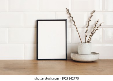 Spring still life. Blank black picture frame mockup on wooden table background. Easter composition with blooming goat willow, pussy willow or great sallow in ceramic vase. White tiles wall background. - Shutterstock ID 1929142775