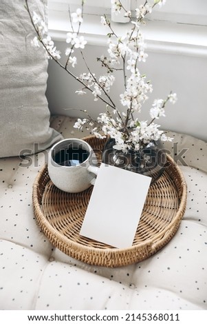 Spring still life composition. Greeting card mockup on wickered tray, cup of coffee. Feminine styled photo. Floral scene with  blurred white cherry tree blossoms on bench near window. Selective focus