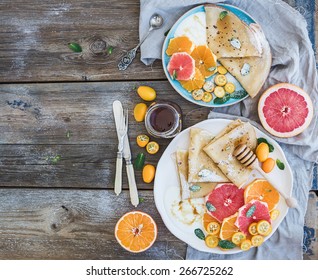 Spring vitamin breakfast set. Thin crepes or pancakes with fresh grapefruit, orange, kumquat, honey, cream and mint leaves over a rustic wood background, top view  - Powered by Shutterstock