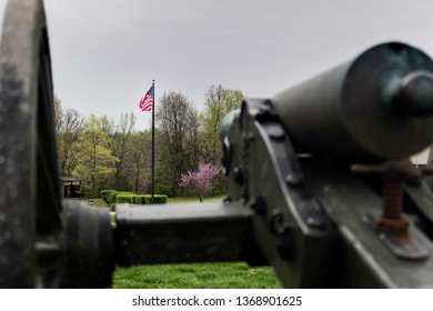 Springfield, MO—April 11, 2019; Bronze Civil War Era Confederate Cannon Points Toward American Flag In Focus At Wilsons Creek National Battlefield In Southwest Missouri In Spring