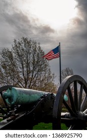 Springfield, MO—April 11, 2019; Bronze Civil War Era Confederate Cannon Points Past American Flag At Wilsons Creek National Battlefield In Southwest Missouri With Sunlight Breaking Through Clouds