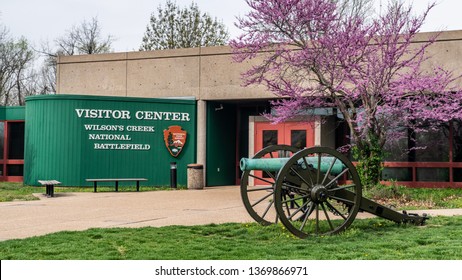 Springfield, MO—April 11, 2019, Bronze Civil War Era Cannon Sits In Front Of The Visitor Center Entrance At Wilsons Creek National Battlefield With Flowering Purple Tree In Spring