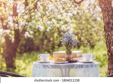 Springtime in garden. Table set with tea cups, boho reuse jar vase with forget me not flowers( Myosotis) and small stack of books. Summer relax readings concept.