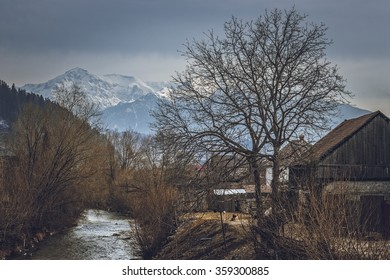 Springtime rural scenery with riverside Romanian traditional village and snowy Bucegi mountains, near Rasnov town, Brasov, county, Transylvania region, Romania.