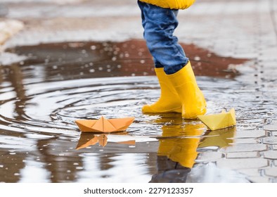 It's springtime. A small child in yellow rubber boots jumps through puddles, plays and launches paper boats on the water.. Photo of spring and autumn holidays. The concept of spring.