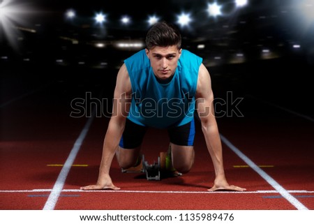 Sprinter leaving starting blocks on the running track in front of big modern stadium with lights. Explosive start.