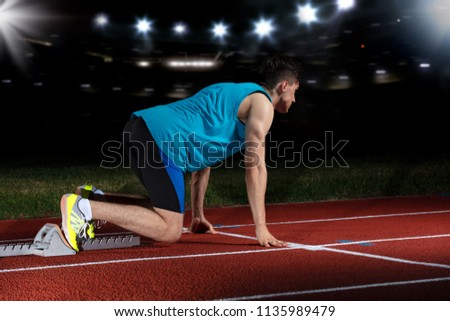 Sprinter leaving starting blocks on the running track in front of big modern stadium with lights. Explosive start.