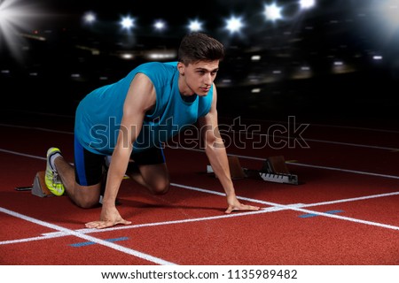Sprinter leaving starting blocks on the running track in front of big modern stadium with lights. Explosive start.