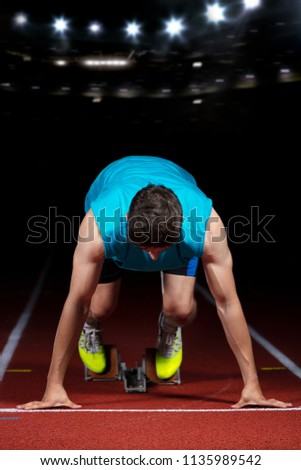 Sprinter leaving starting blocks on the running track in front of big modern stadium with lights. Explosive start.