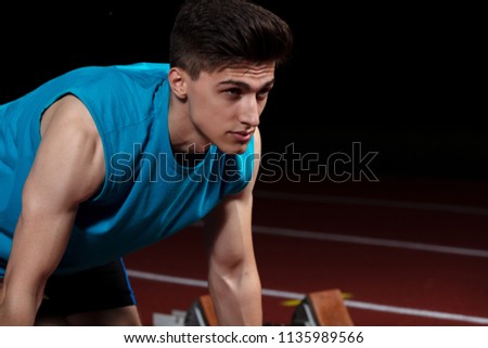 Sprinter leaving starting blocks on the running track in front of big modern stadium with lights. Explosive start.