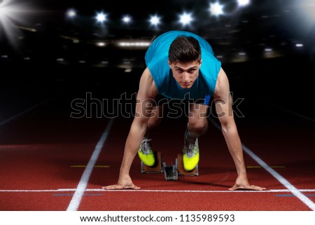 Sprinter leaving starting blocks on the running track in front of big modern stadium with lights. Explosive start.