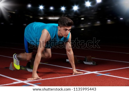 Sprinter leaving starting blocks on the running track in front of big modern stadium with lights. Explosive start.
