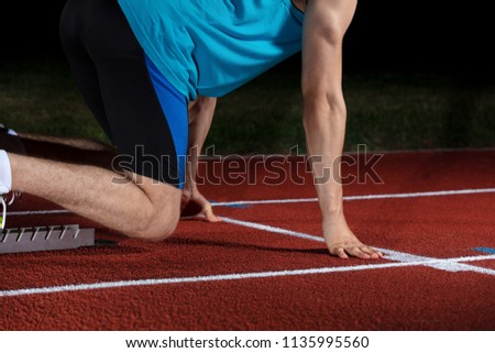Sprinter leaving starting blocks on the running track in front of big modern stadium with lights. Explosive start.