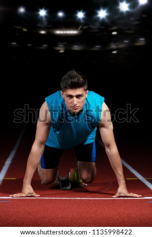Sprinter leaving starting blocks on the running track in front of big modern stadium with lights. Explosive start.