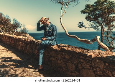 A spruce black guy in a stylish blue costume is holding his straw hat from the wind while sitting on a stone fence outdoors next to the ocean coast surrounded by trees on a warm sunny day