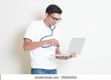 Spyware Or Malware Concept. Indian Computer Engineer Diagnose Virus Infected Pc With Stethoscope. Asian Man Standing On Plain Background With Shadow And Copy Space. 