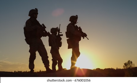 Squad of Three Fully Equipped and Armed Soldiers Standing on Hill in Desert Environment in Sunset Light.