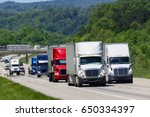 A squadron of eighteen-wheelers lead the way down an interstate highway in eastern Tennessee.  Heat waves rising from the pavement give a shimmering effect to vehicles and forest in the background.