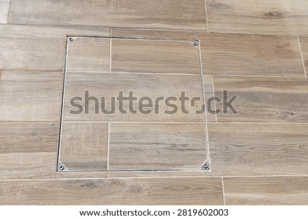 A square access hatch is discreetly set into a floor covered with modern wood-look ceramic tiles, featuring visible screw heads