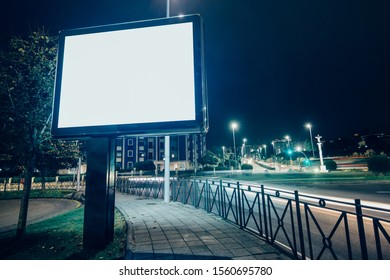 Square Billboard Mockup With Blank Canvas Screen At Night In The City With A Roundabout With Moving Traffic In The Background.