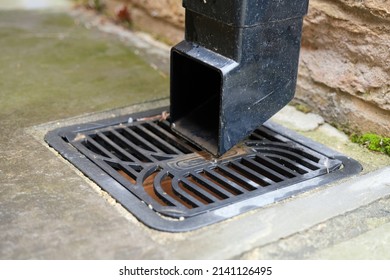 Square black plastic drainage downpipe with a down spout shoe at the end of it. Fixed to a brick wall. Leading to a black plastic square gulley grid type drainage cover. Day outdoors. Close up.