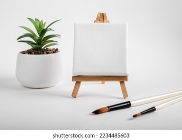 Square canvas on wood mini easel, blank mock up on wood tripod, paintbrushes and potted plant composition. High quality photo - Powered by Shutterstock