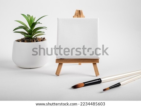 Square canvas on wood mini easel, blank mock up on wood tripod, paintbrushes and potted plant composition. High quality photo