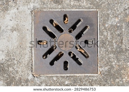 Square cast iron drainage grate with a radial slot pattern installed in a textured concrete floor surface to collect surface water runoff.